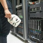 Osoba z maską anonimowego hakera stojąca przy serwerach komputerowych